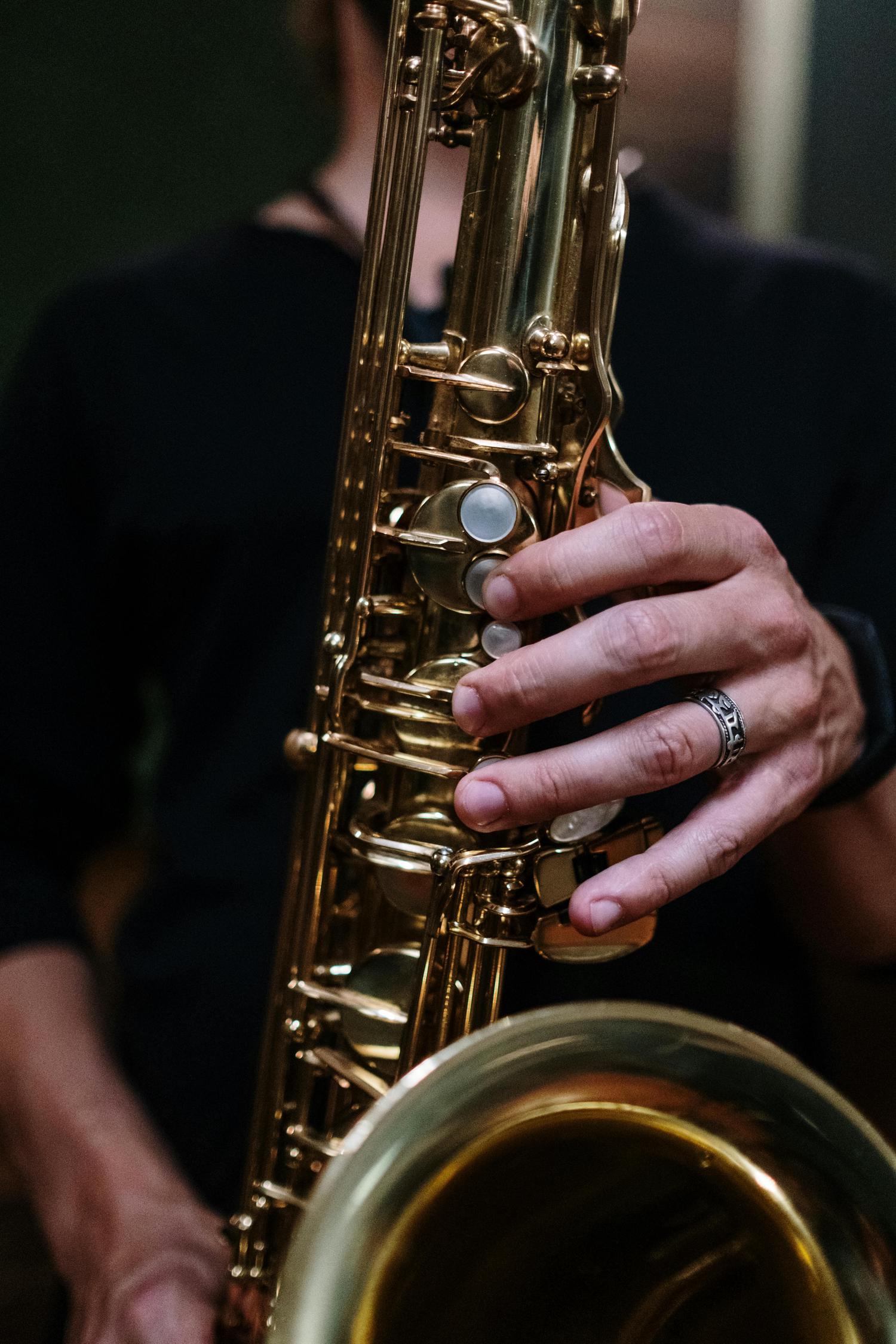 A close-up shows a person holding a golden saxophone with their left hand, pressing the keys. The person wears a ring on their left ring finger, and is dressed in black.