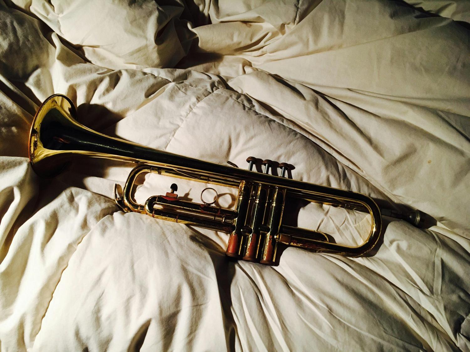 A gold-colored trumpet lies on a rumpled, white comforter. The instrument is positioned diagonally, with its bell facing the upper-left corner.