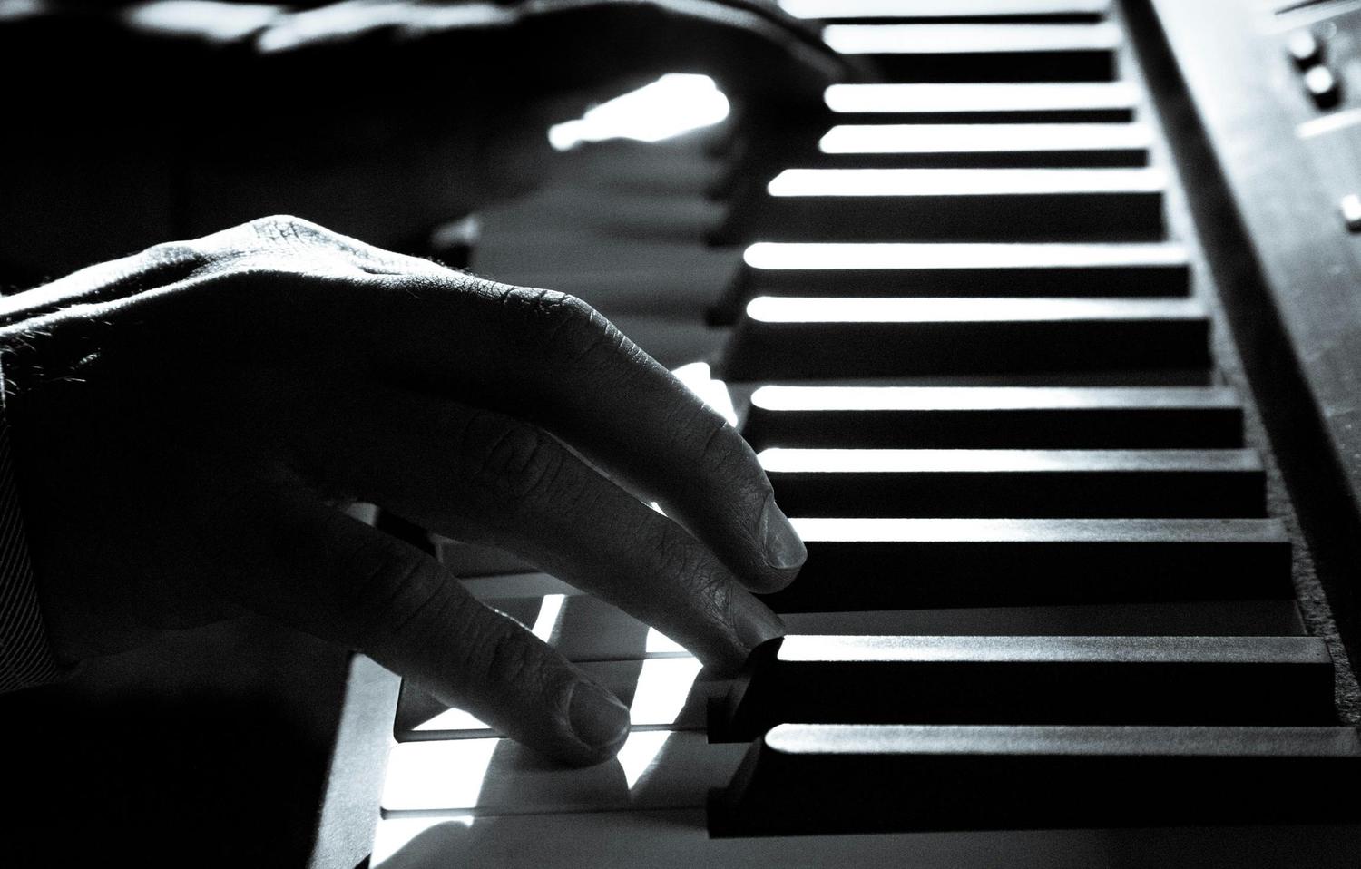 Close-up, black and white shot of a person's hands playing a piano keyboard. Light streams across the keys as fingers depress one of the black keys.