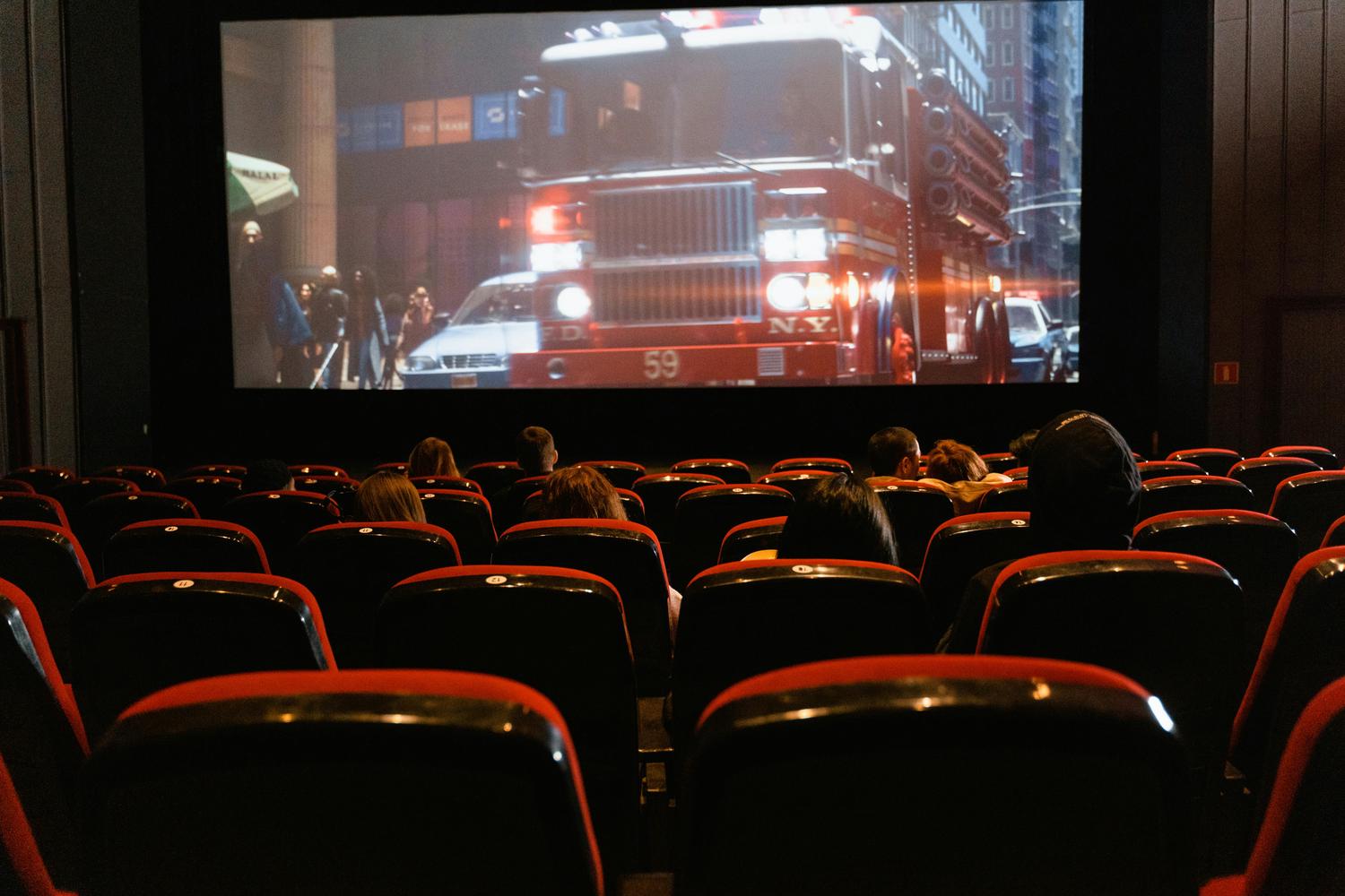 View from the back of a darkened movie theater showing rows of mostly filled red seats facing a large screen. Onscreen is a film depicting a New York City fire truck.