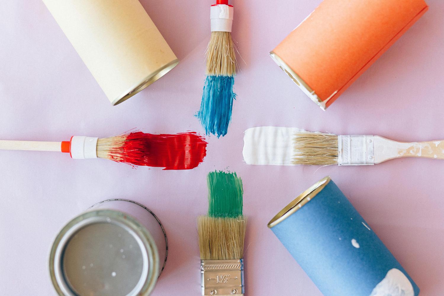 Overhead shot of an art setup, featuring paintbrushes with strokes of red, blue, white, and green paint arranged with corresponding open paint cans on a light pink background. The composition suggests an artistic or DIY painting project.