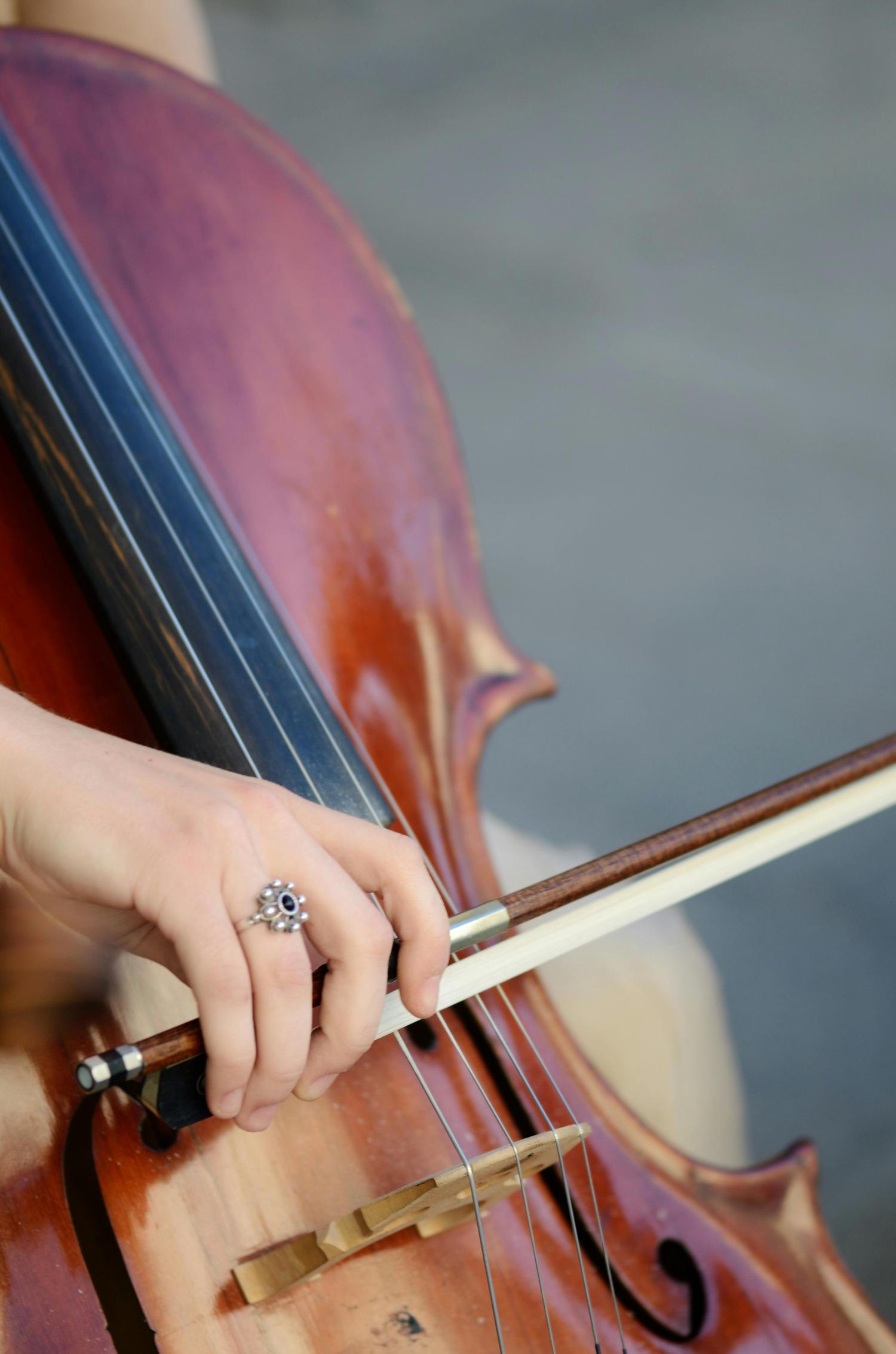 Close-up of a hand with an ornate ring holding a bow and playing a cello. The instrument's warm, reddish-brown wood and strings are clearly visible.