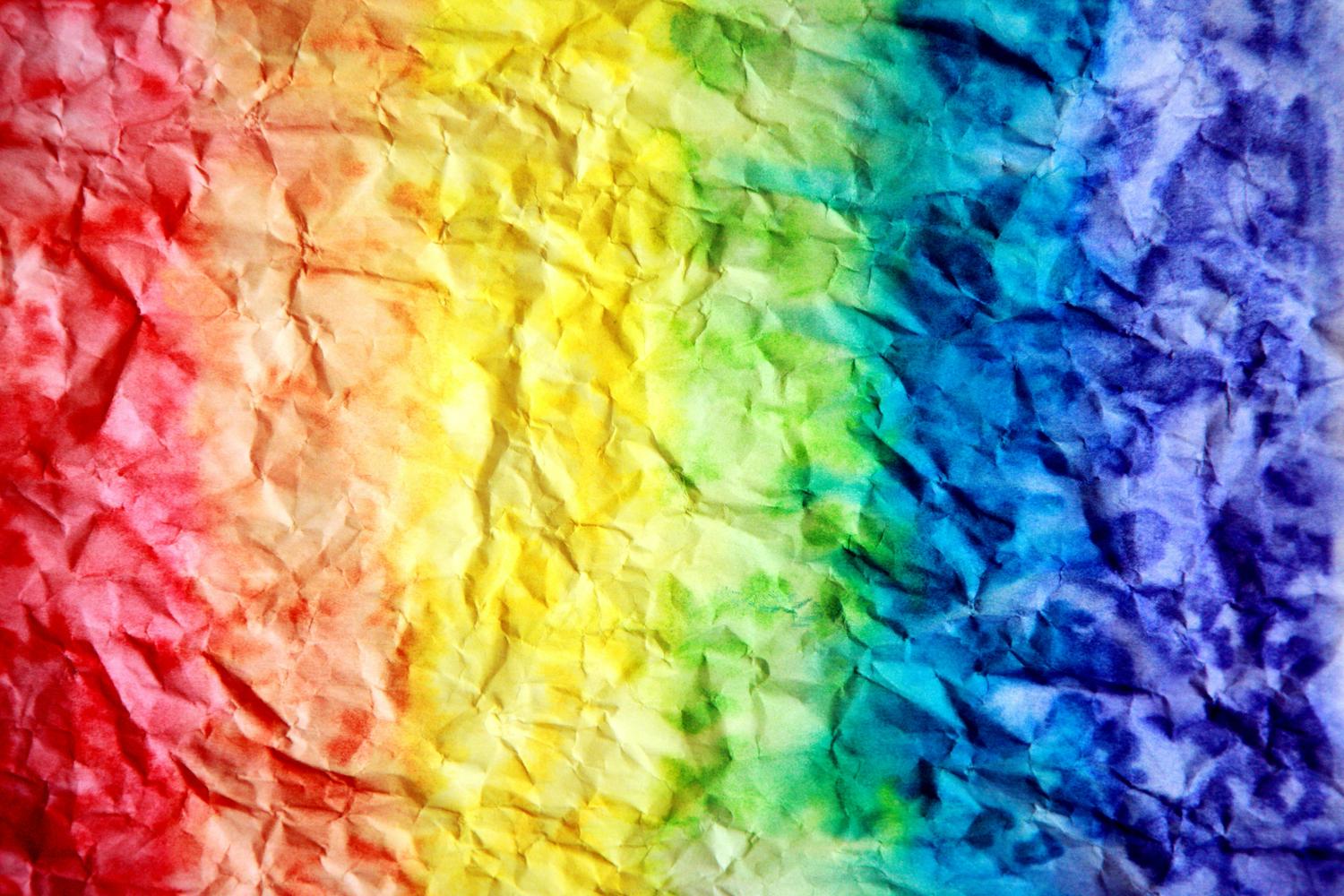 Crumpled paper is dyed with a rainbow gradient, transitioning from red to yellow to green to blue and purple. The texture of the paper creates variations in color intensity across the surface.