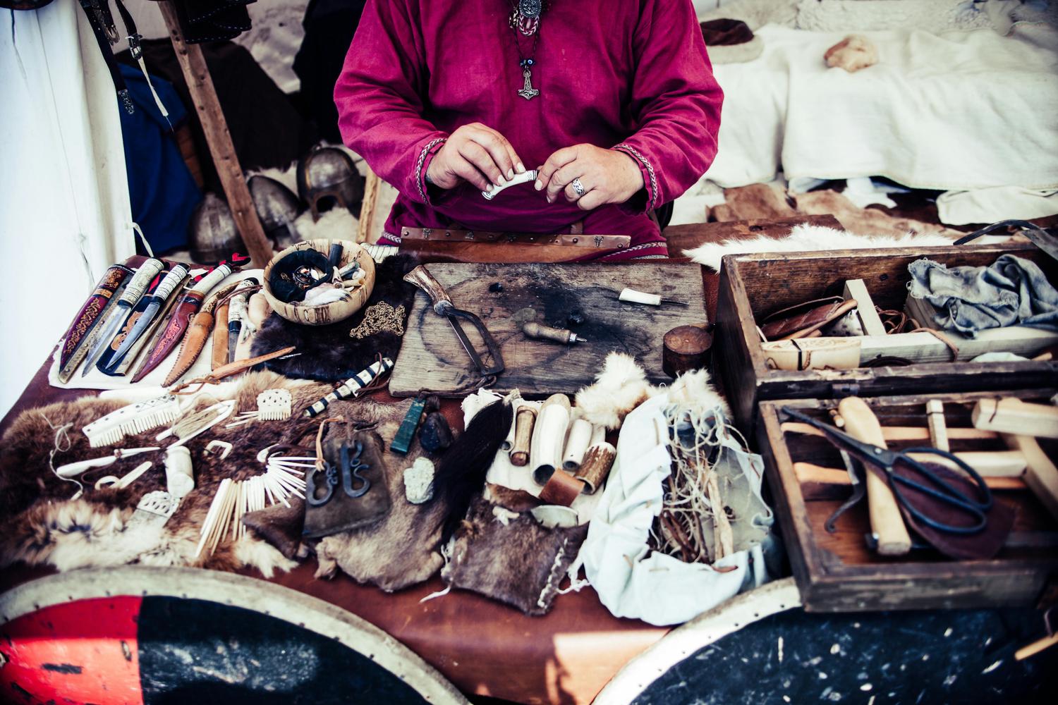 A person in a red tunic is crafting with bone at a table laden with tools, animal hides, knives, and wooden boxes, suggesting a historical reenactment or artisan's workshop. The scene is illuminated with a slightly muted, vintage-style filter.