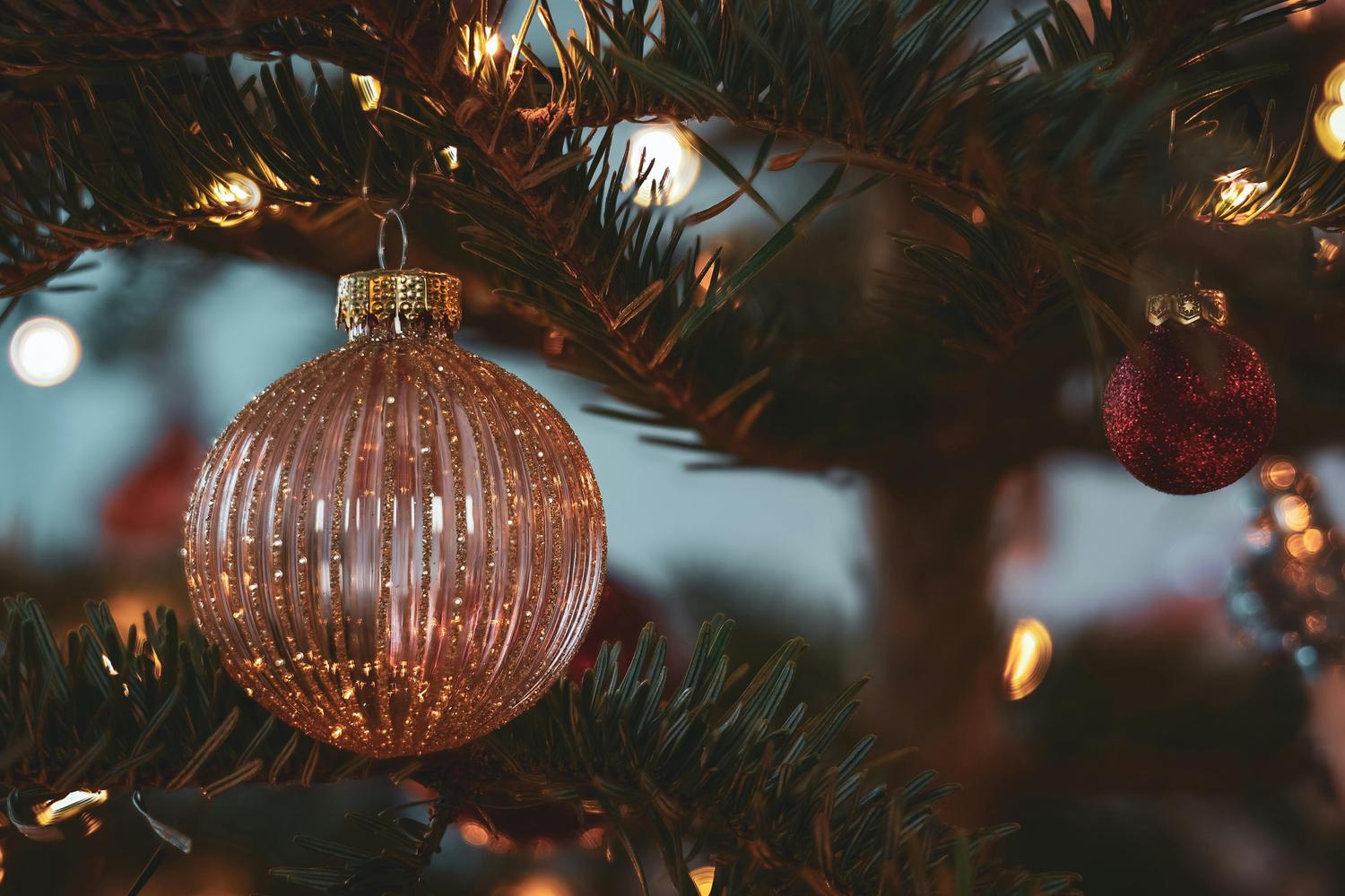 A close-up view of a Christmas tree with a rose gold-colored, vertically ridged ornament in the foreground and a smaller, red ornament visible in the background, with soft focus lights interspersed amongst the green pine needles. The scene evokes a warm, festive ambiance.