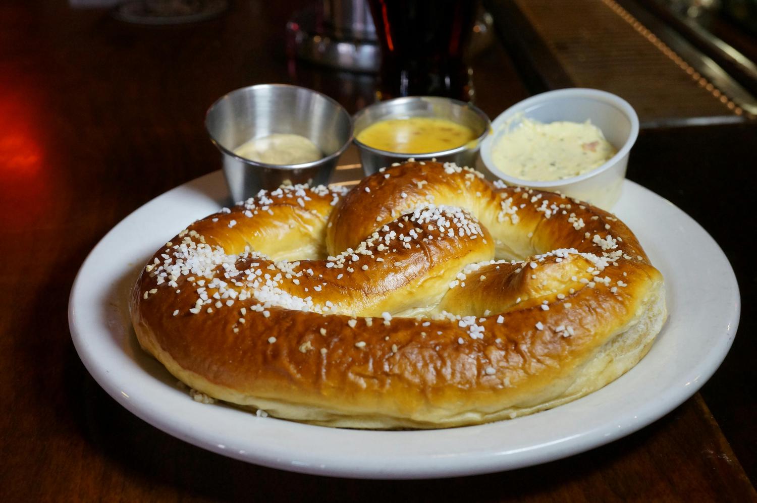 A large, golden-brown pretzel sprinkled with salt sits on a white plate, accompanied by three dipping sauces in small containers. The sauces appear to be honey mustard, cheese sauce, and herbed butter.