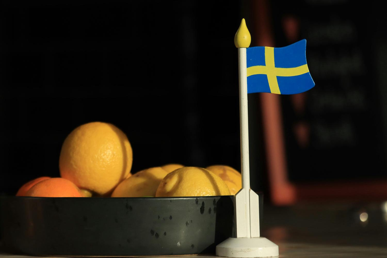 A bowl of lemons and oranges sits next to a small Swedish flag on a tabletop. The background is dark and out of focus.