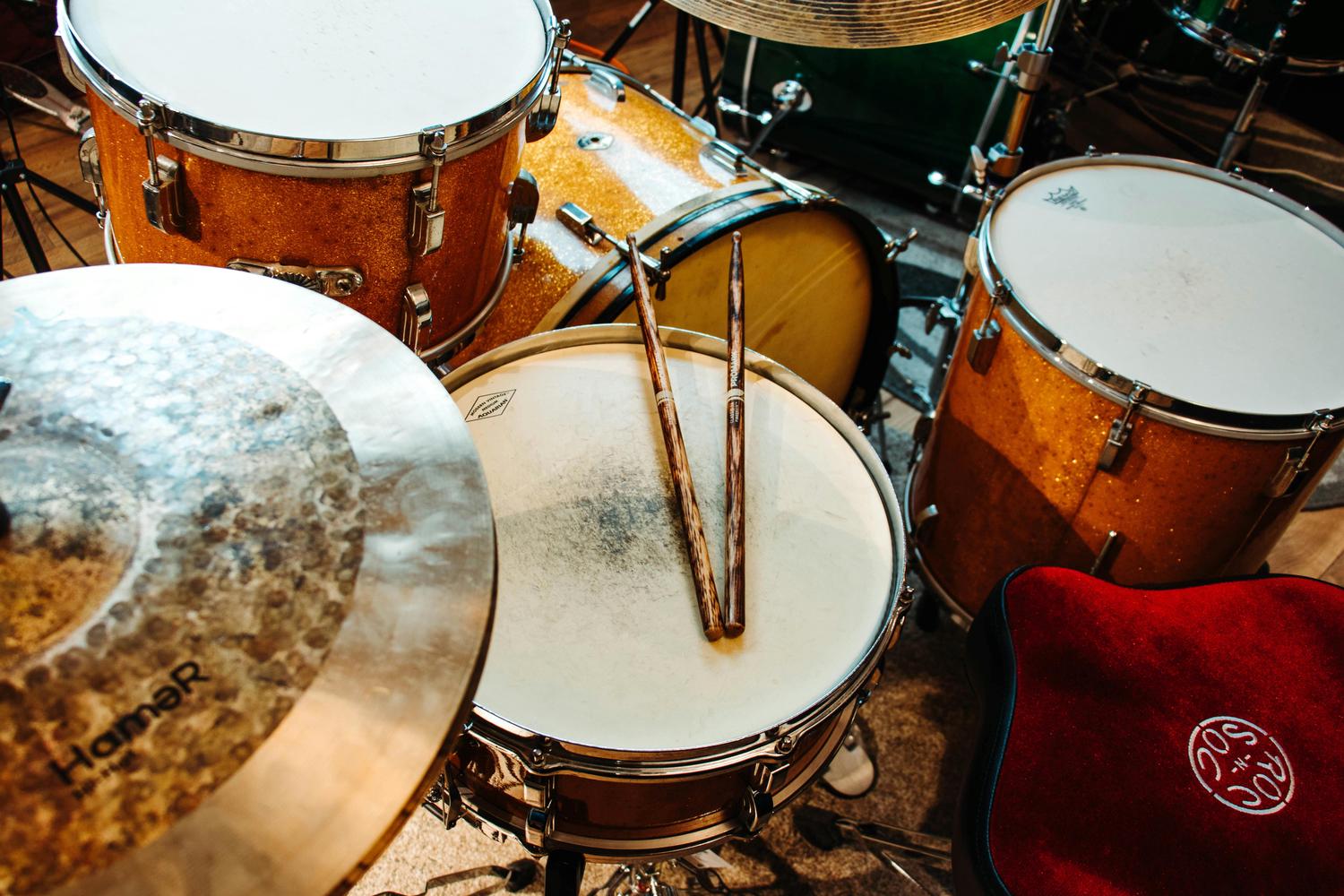 A close-up view of a drum set with brown and gold glitter drums and cymbals, featuring a pair of wooden drumsticks resting on a snare drum. A red drum throne is partially visible in the lower right corner.