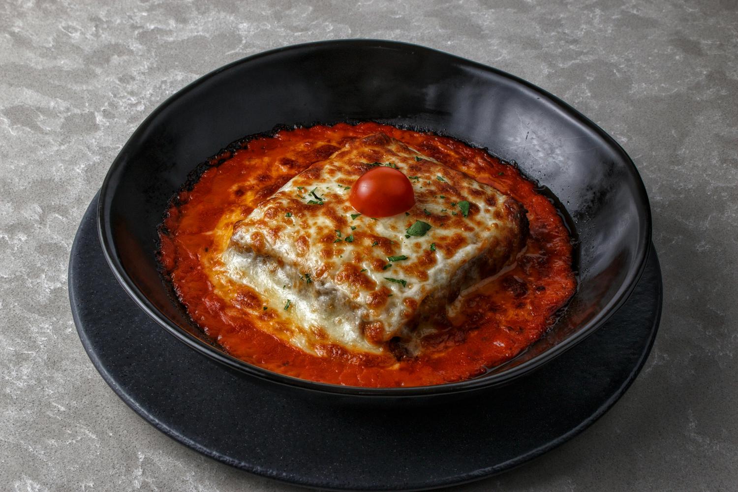 A close-up shows lasagna topped with melted cheese and a cherry tomato, sitting in a black bowl with red sauce. The bowl is on a black plate against a gray countertop.