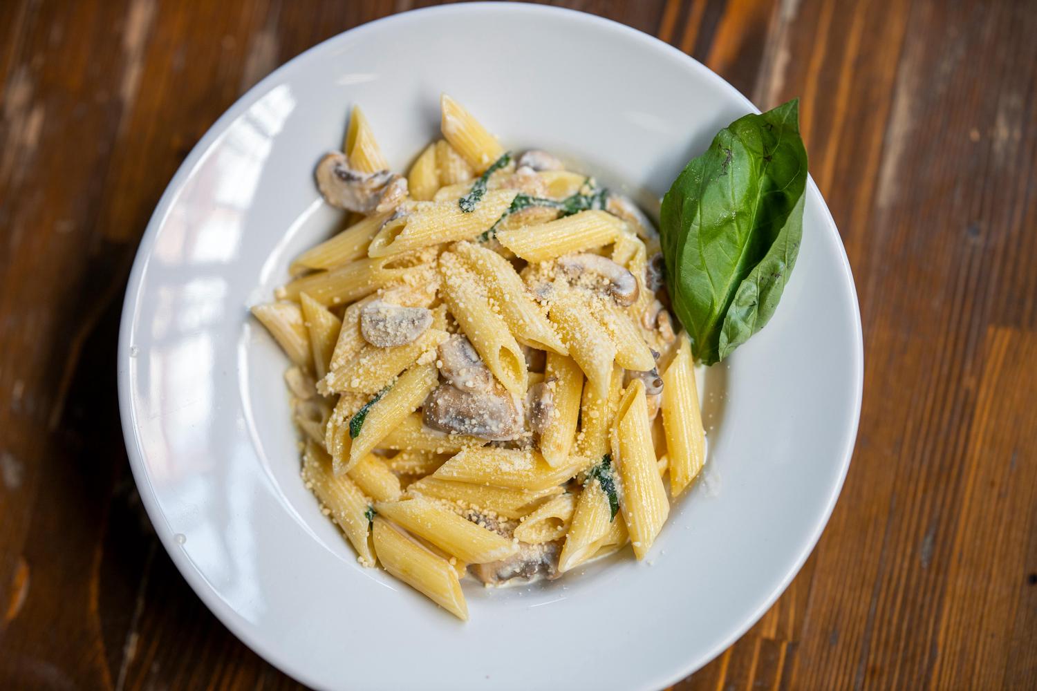 A plate of penne pasta with creamy sauce, mushrooms, and spinach is garnished with parmesan cheese and a basil leaf, sitting on a wooden surface. The dish is presented from an overhead perspective, highlighting its textures and ingredients.