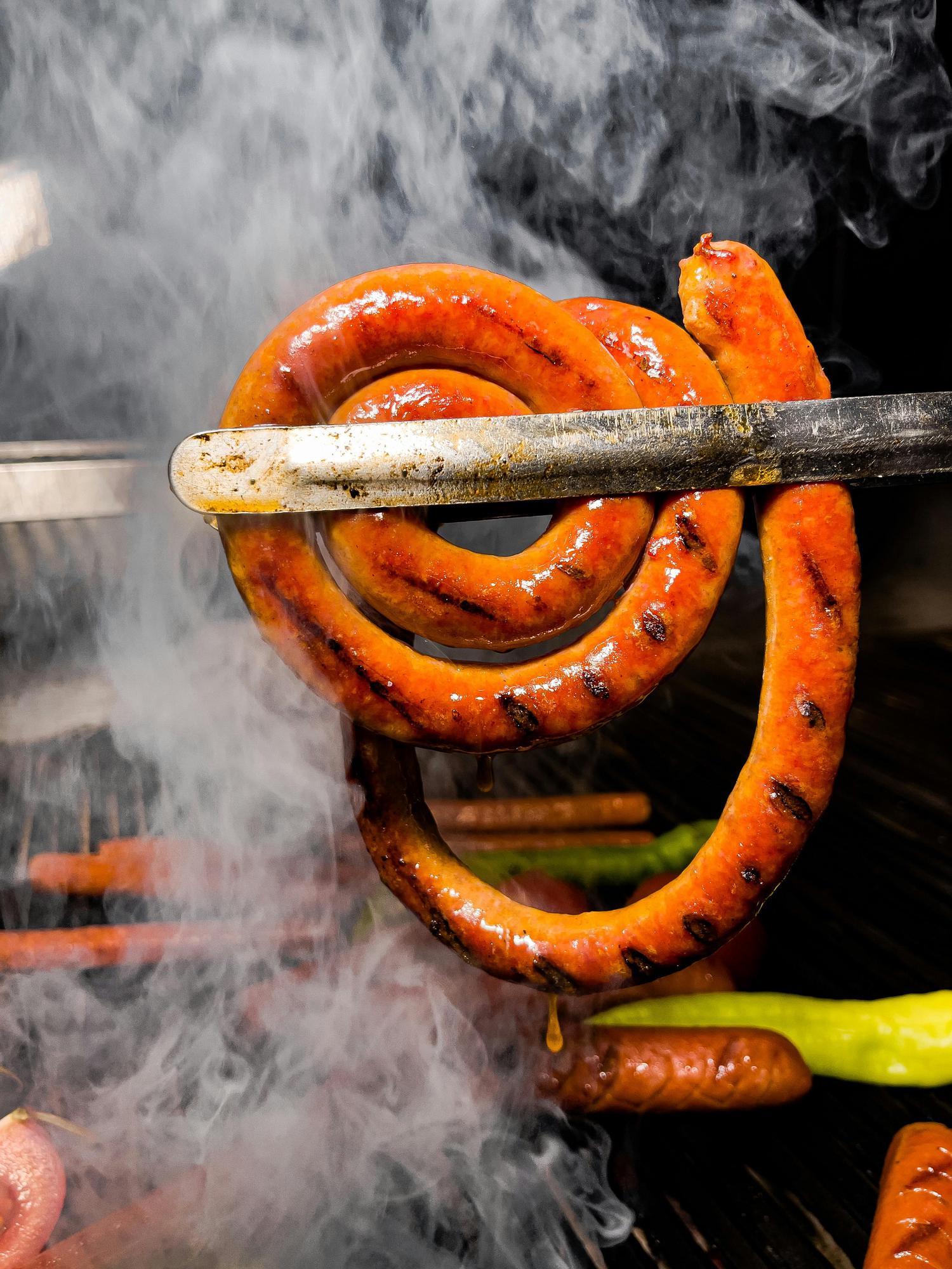 A long, coiled sausage is held by metal tongs, surrounded by smoke from a grill. Other sausages and vegetables are visible on the grill in the background.