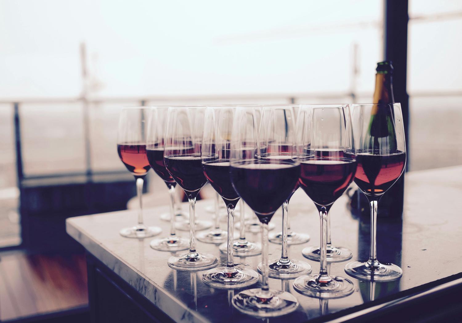 Multiple wine glasses filled with red wine stand in neat rows on a table, with a champagne bottle visible in the background. The scene suggests an event or celebration.