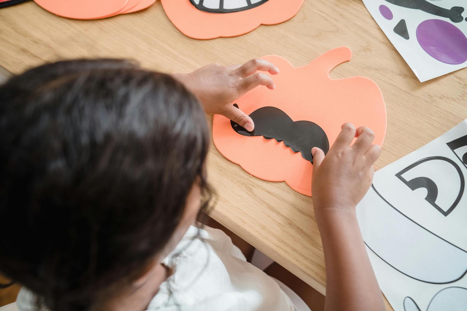 A child is shown from above, attaching a black, scalloped mouth to an orange pumpkin cutout on a wooden table, likely creating Halloween decorations. Additional pumpkin cutouts and other Halloween-themed shapes are visible nearby.