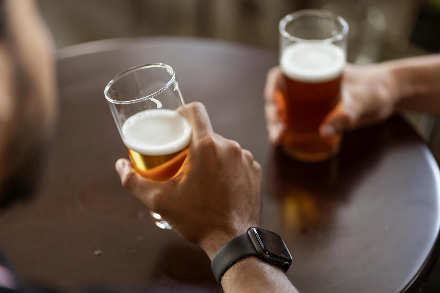 Two people are holding glasses of golden beer with foamy heads at a dark brown table. One person has a smartwatch on their wrist.
