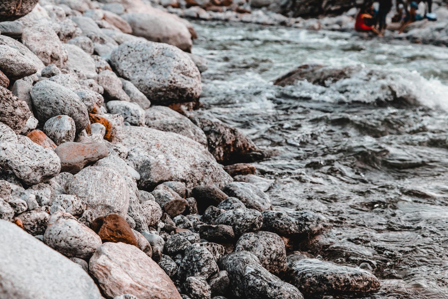 A fast-moving river flows past a rocky bank covered in grey and white speckled stones. In the distance, a blurred group of people stands in the water.