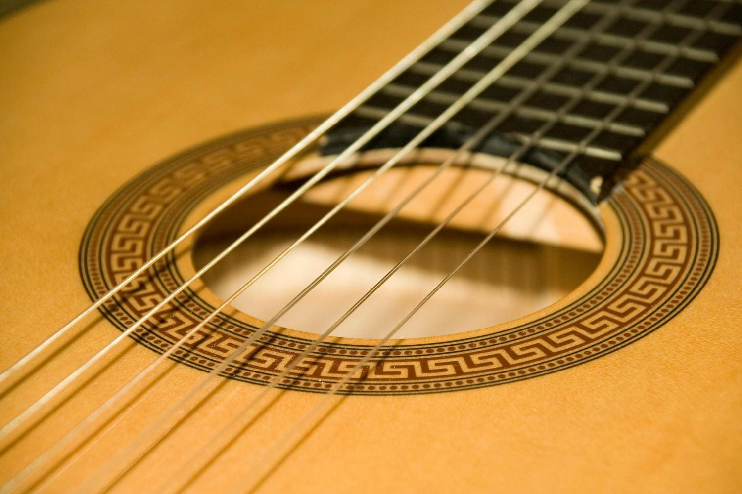 Close-up of an acoustic guitar, focusing on the sound hole with its decorative patterned border and the guitar strings. The fretboard is visible in the background.