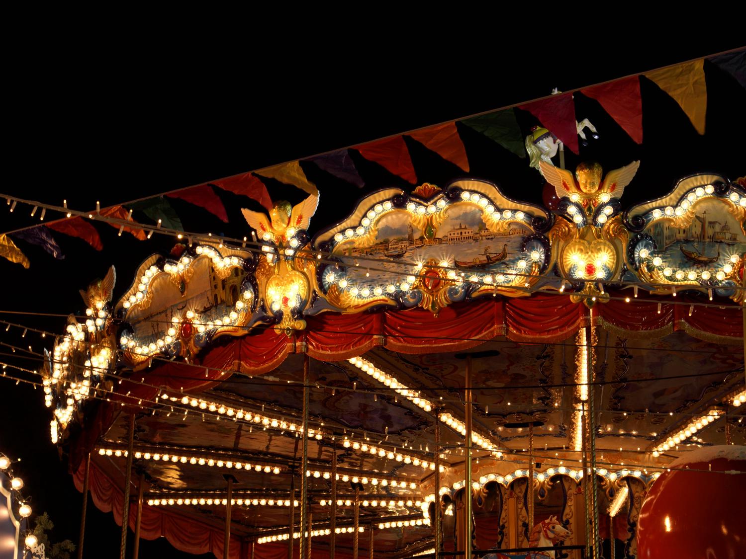 A carousel at night is brightly lit with strings of lights; the top is ornamented with cherubs and painted scenes, and a string of colorful flags is above. The foreground shows the inner workings of the carousel and some of the painted horses.