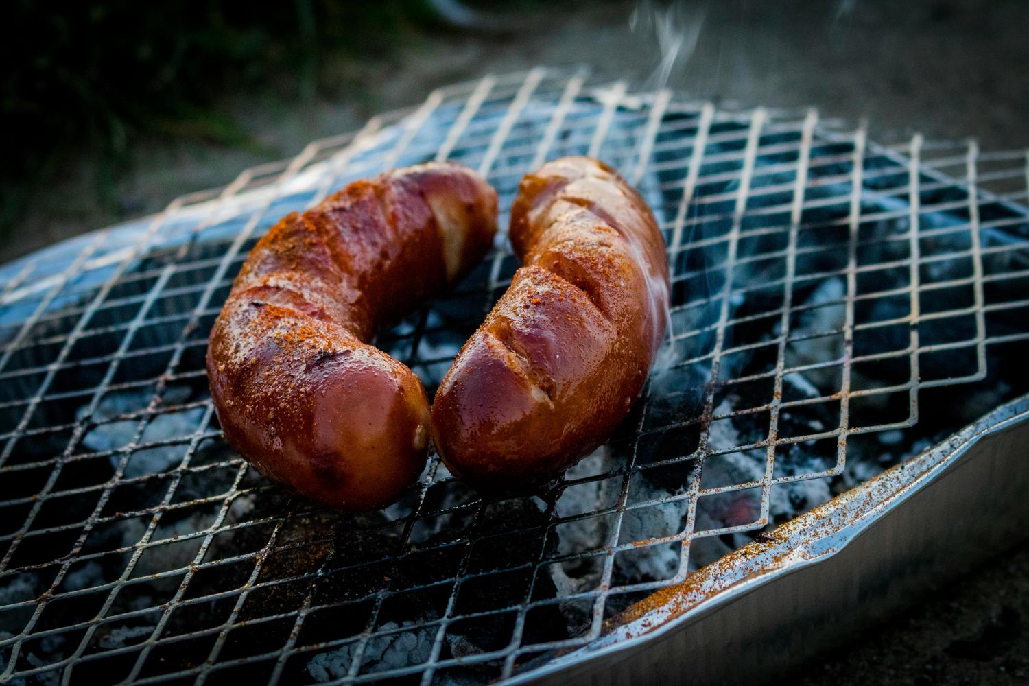 Two grilled sausages sit on a metal grate, with wisps of smoke rising around them. The grill sits above a bed of ash.