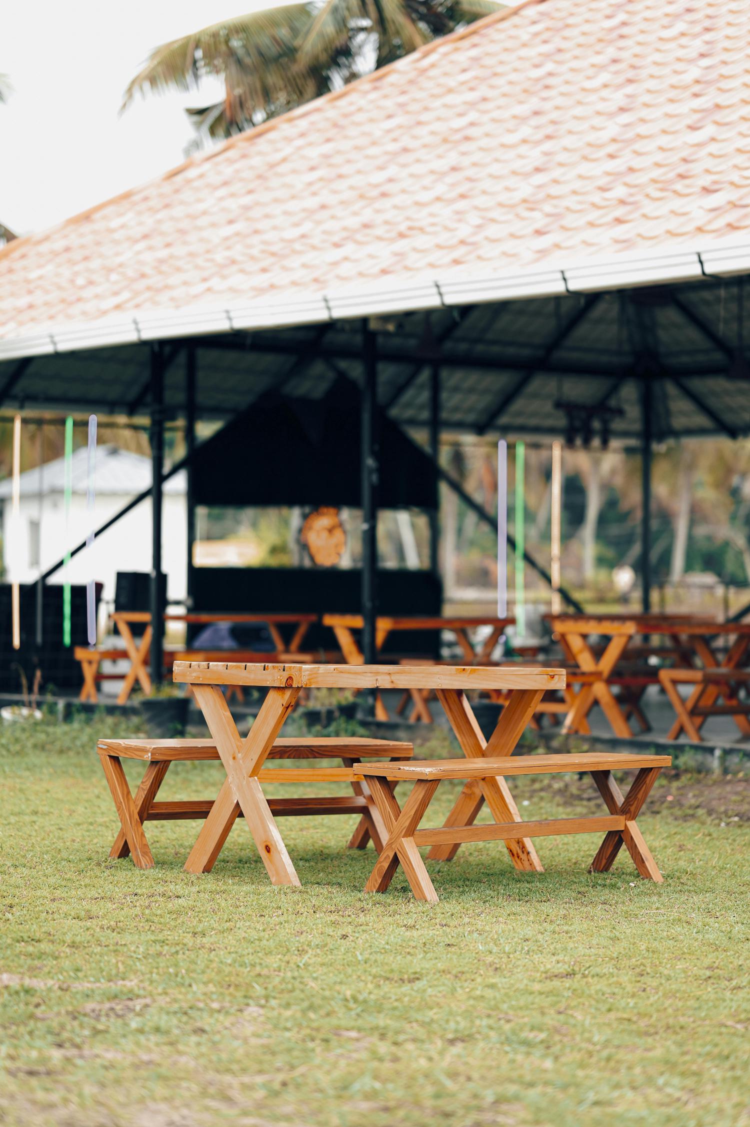 A picnic table made of light-colored wood sits on a lawn in front of a covered pavilion, suggesting an outdoor dining or recreational area. Additional tables are visible inside the pavilion.