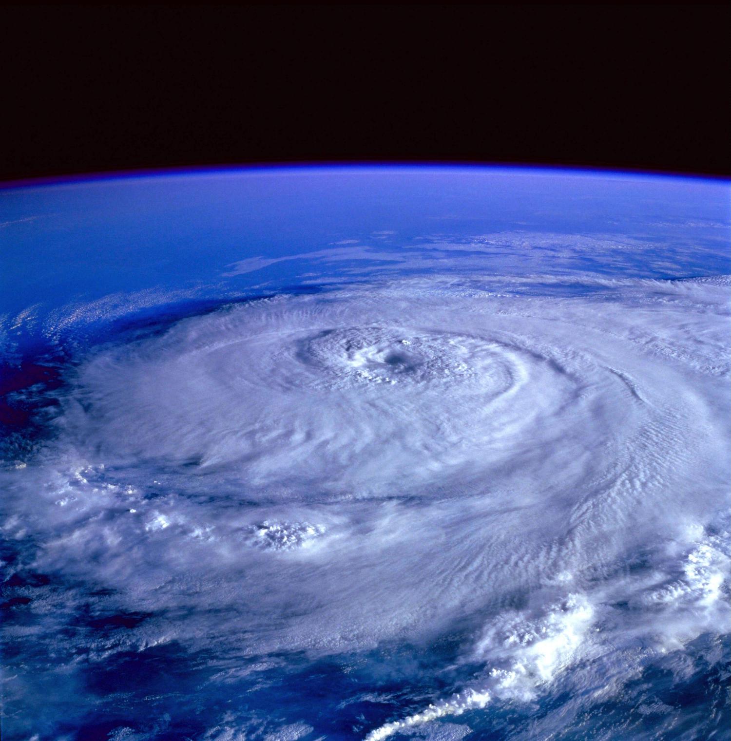 A view of a large hurricane swirling over the ocean, taken from space, with the blue curve of the Earth visible and black space above. The eye of the storm is clearly visible within the spiraling bands of clouds.