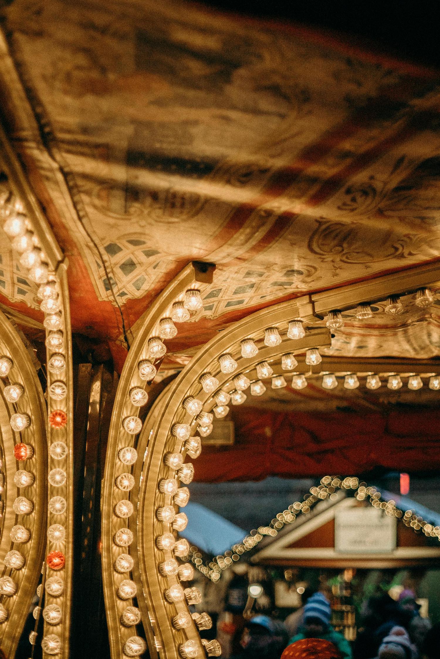 Close-up of a vintage carousel with golden supports illuminated by strings of lights, topped by a painted fabric canopy. In the background, blurred market stalls and people suggest a festive, outdoor atmosphere.