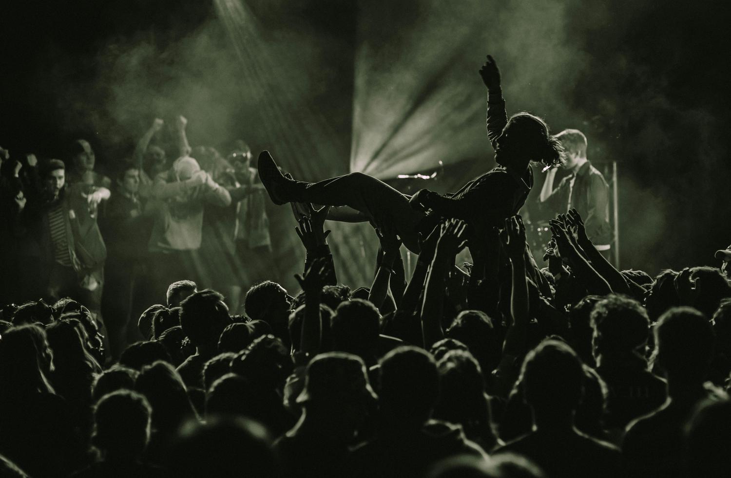 A silhouetted crowd scene at a concert, with a person crowd surfing and raising their arm in the air, lit by a spotlight from above. Many hands reach up from the crowd to support the person.