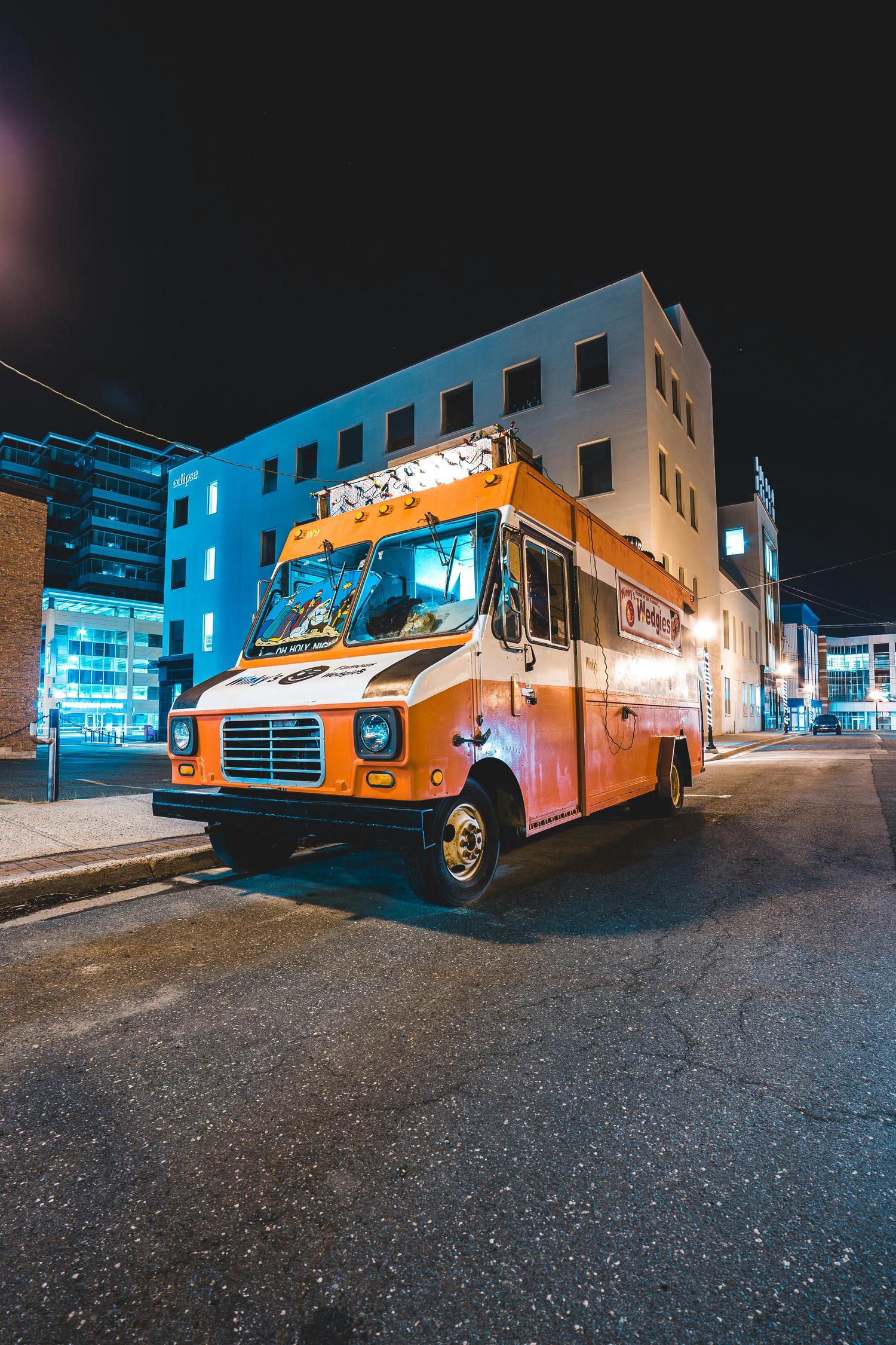 An orange and white food truck, illuminated from within, is parked on a city street at night, with tall buildings in the background. The truck features holiday-themed decorations above the windshield and signage for 