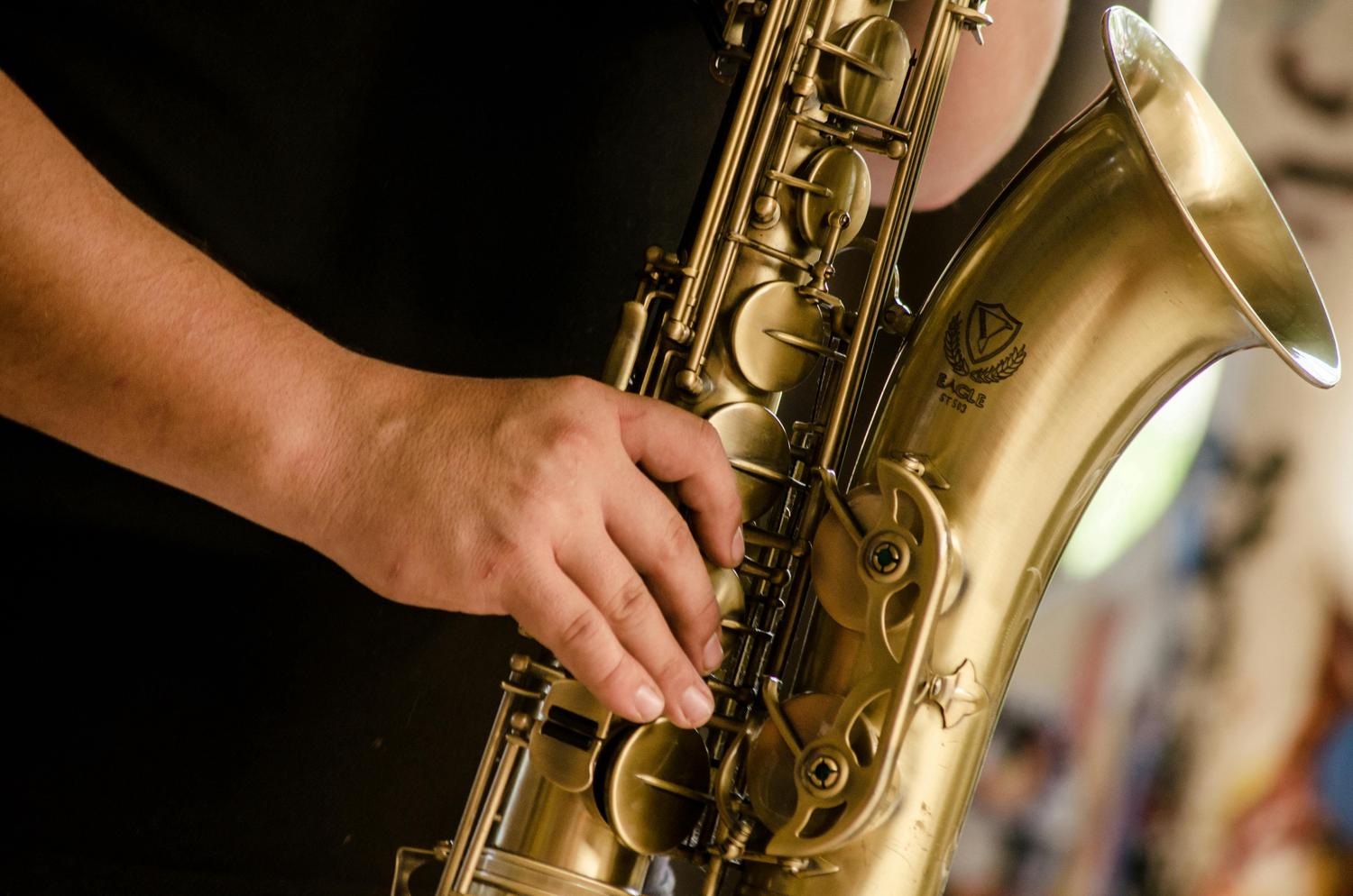 Close-up view of a hand pressing the keys of a shiny, brass-colored saxophone. The instrument's bell and intricate key mechanisms are visible.
