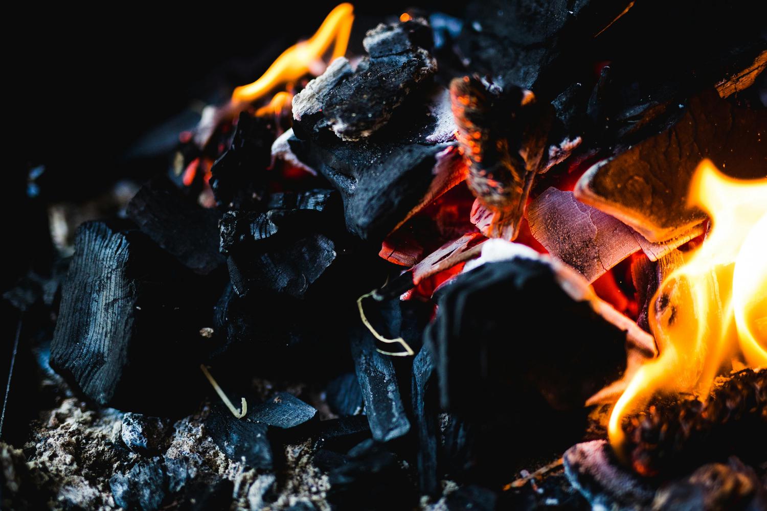 Burning coal emits bright orange and yellow flames in a close-up shot. The black charcoal is in various shapes and sizes, resting on a bed of ash.