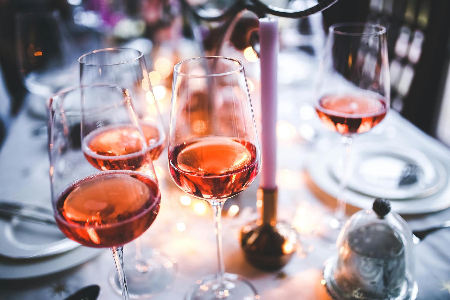 A table is set for a formal event, featuring wine glasses filled with rosé wine, a tall pink candle, plates, and silverware. The scene is illuminated by soft, warm lighting, creating a festive and elegant atmosphere.