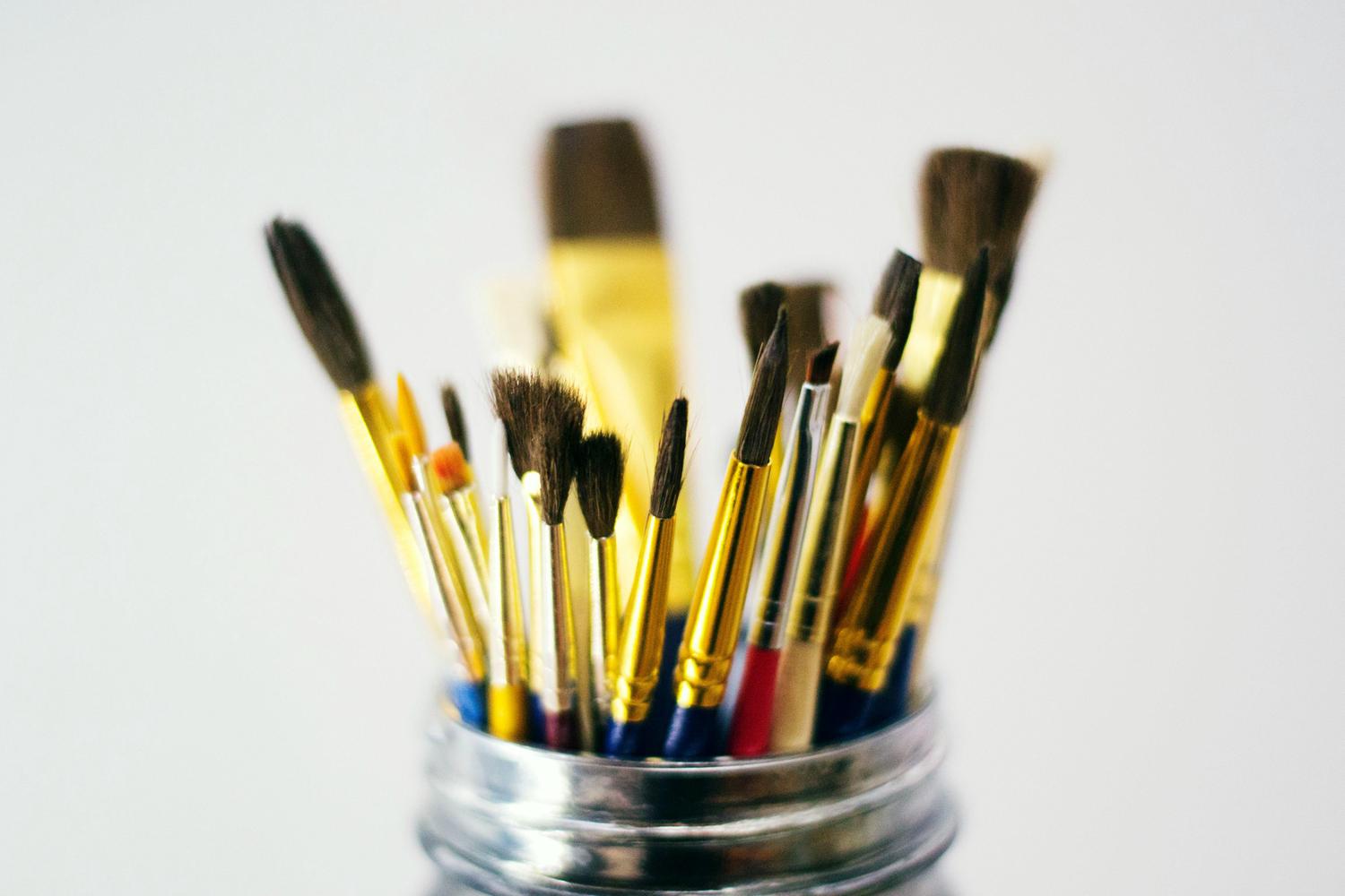 A variety of paintbrushes, some with gold handles, are arranged inside a silver jar, against a plain white backdrop. The brush tips vary in size and shape.