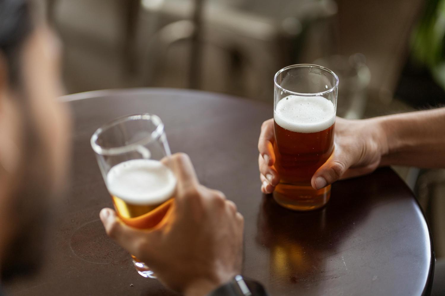 Two people hold glasses of beer at a brown table in a bar or restaurant setting. The beers have white frothy heads, and the glass on the right contains a darker amber beer.