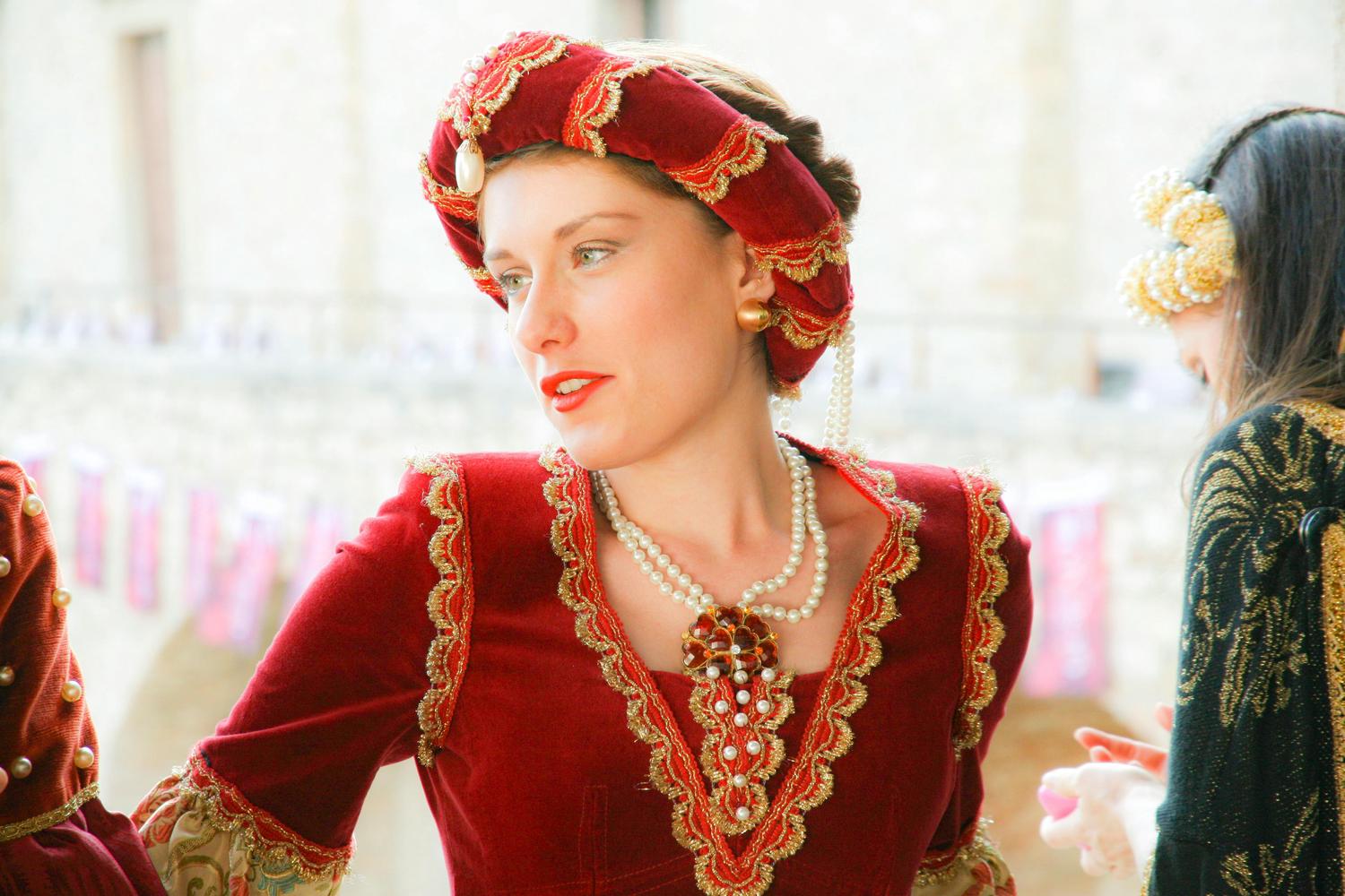 A woman in a red velvet dress and headdress with gold trim and pearl accents looks to the side. The woman is also wearing a pearl necklace and earrings.