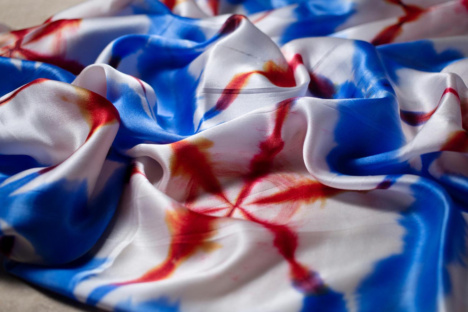 A close-up shows folds of a shiny, white silk scarf with abstract red and blue patterns resembling tie-dye or watercolor washes. The fabric's texture is soft and flowing, with highlights emphasizing the sheen of the silk.