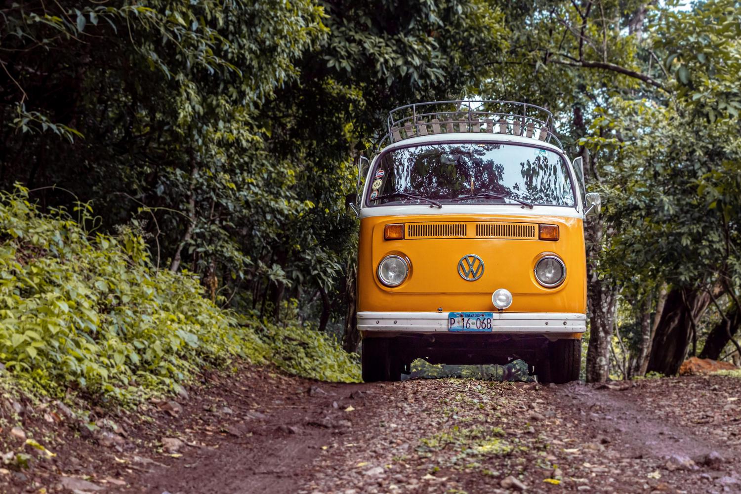 A vintage yellow Volkswagen van faces forward on a dirt road surrounded by dense green foliage. The van features a white roof, a roof rack, and a Salvadoran license plate.