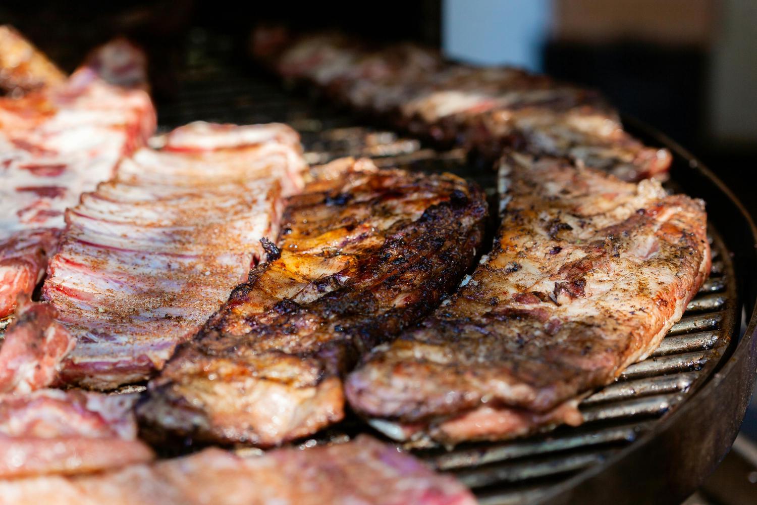 Several racks of ribs are arranged on a metal grill, some appearing raw with seasoning and others showing dark, cooked grill marks. The focus is on the contrast between the uncooked and cooked meat.