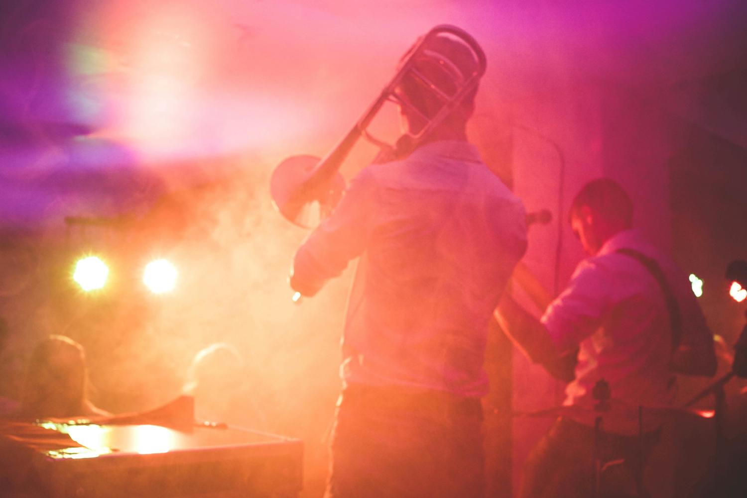A musician plays the trombone on stage, bathed in pink and orange light with hazy smoke effects. Other band members and instruments are visible in the background.