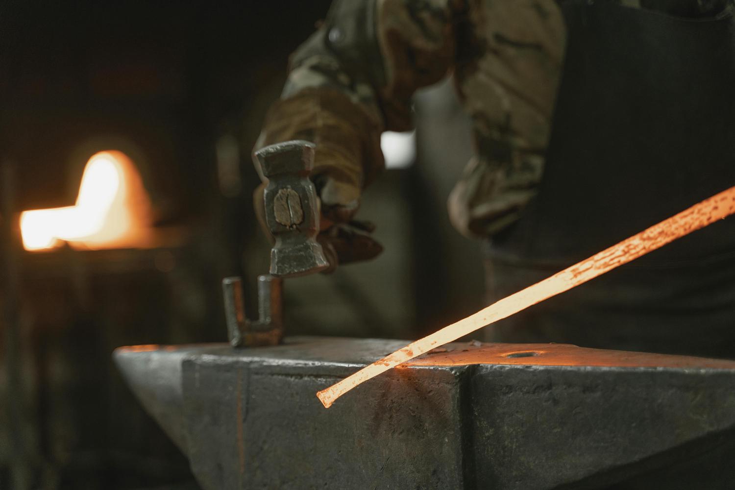 A blacksmith in protective gear hammers a glowing-hot metal rod on an anvil, with the forge fire visible in the background. The image conveys the heat and physical effort involved in metalworking.