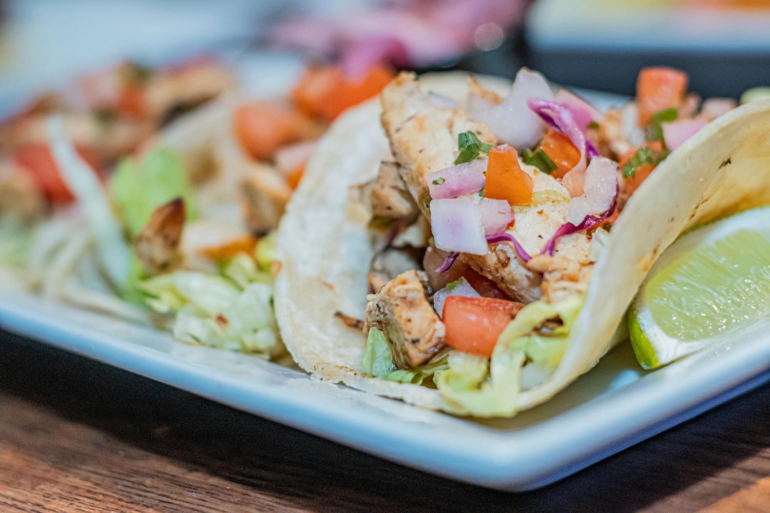 Three fish tacos, filled with diced grilled fish, onions, cabbage, and tomatoes, are arranged on a white rectangular plate alongside a lime wedge. The tacos are on a wooden surface.