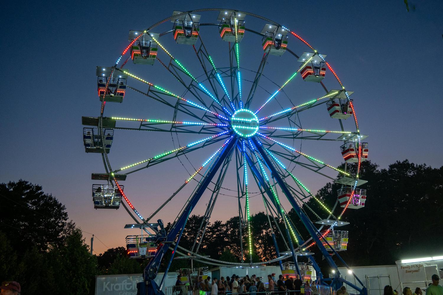 At dusk, a Ferris wheel is illuminated with colorful lights, featuring enclosed cars and riders. Below, a line of people waits near a 