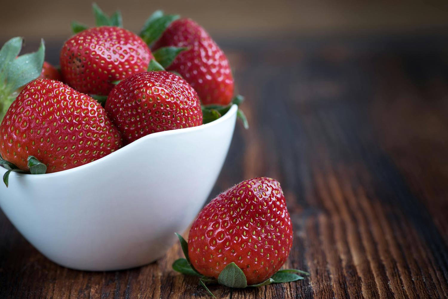 A white bowl filled with ripe, red strawberries sits on a dark wooden surface, with one strawberry positioned outside the bowl. The strawberries have green stems and tiny seeds visible on their surface.