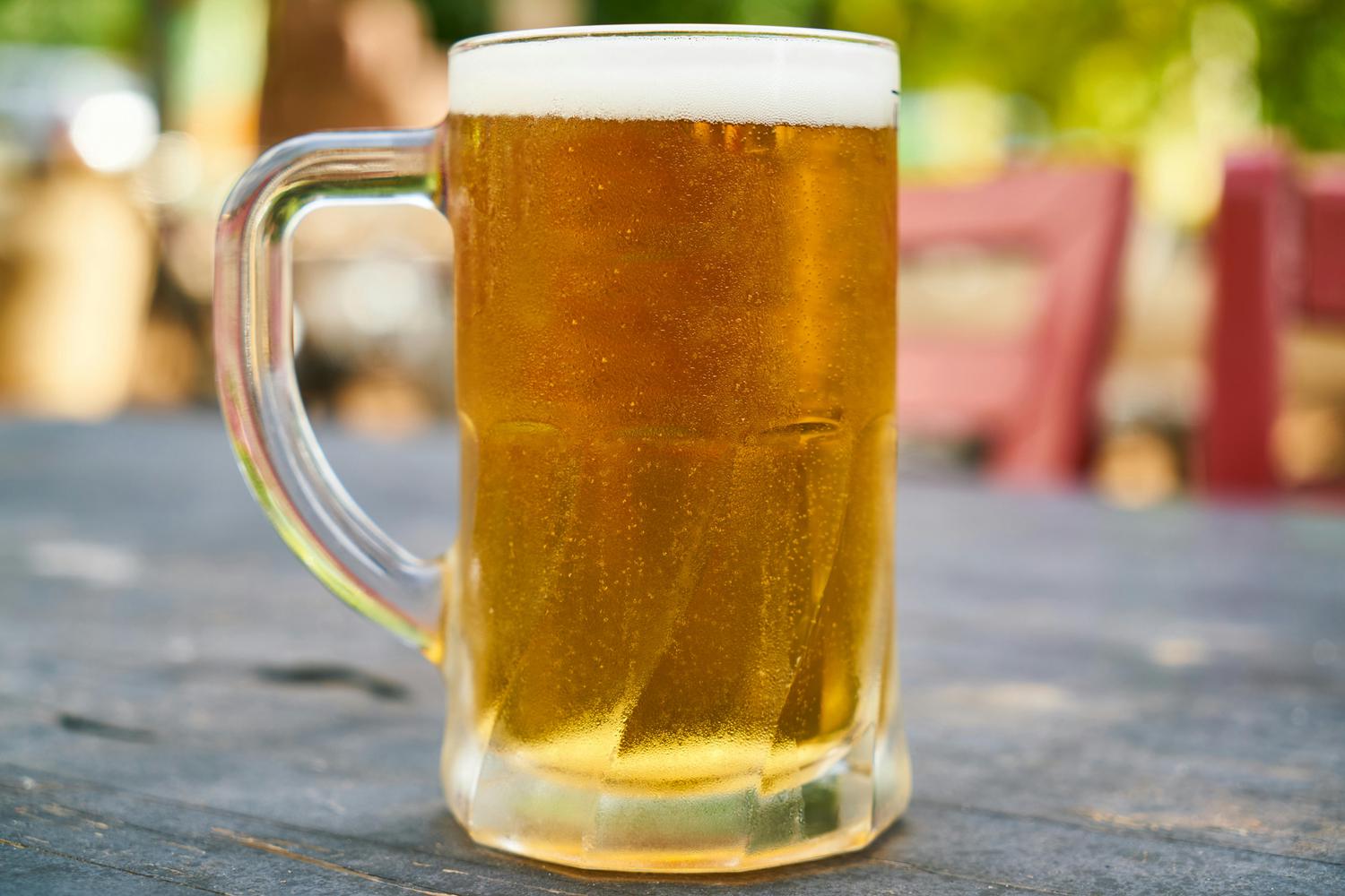 A frosty mug of golden beer with a thick white head sits on a weathered wooden table. The background is blurred with hints of foliage and chairs.