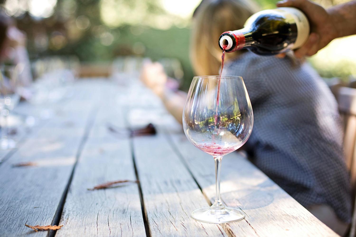 Red wine is being poured from a dark bottle into a clear wine glass on a weathered wooden table, with blurred figures suggesting an outdoor gathering in the background. The focus is on the wine filling the glass.