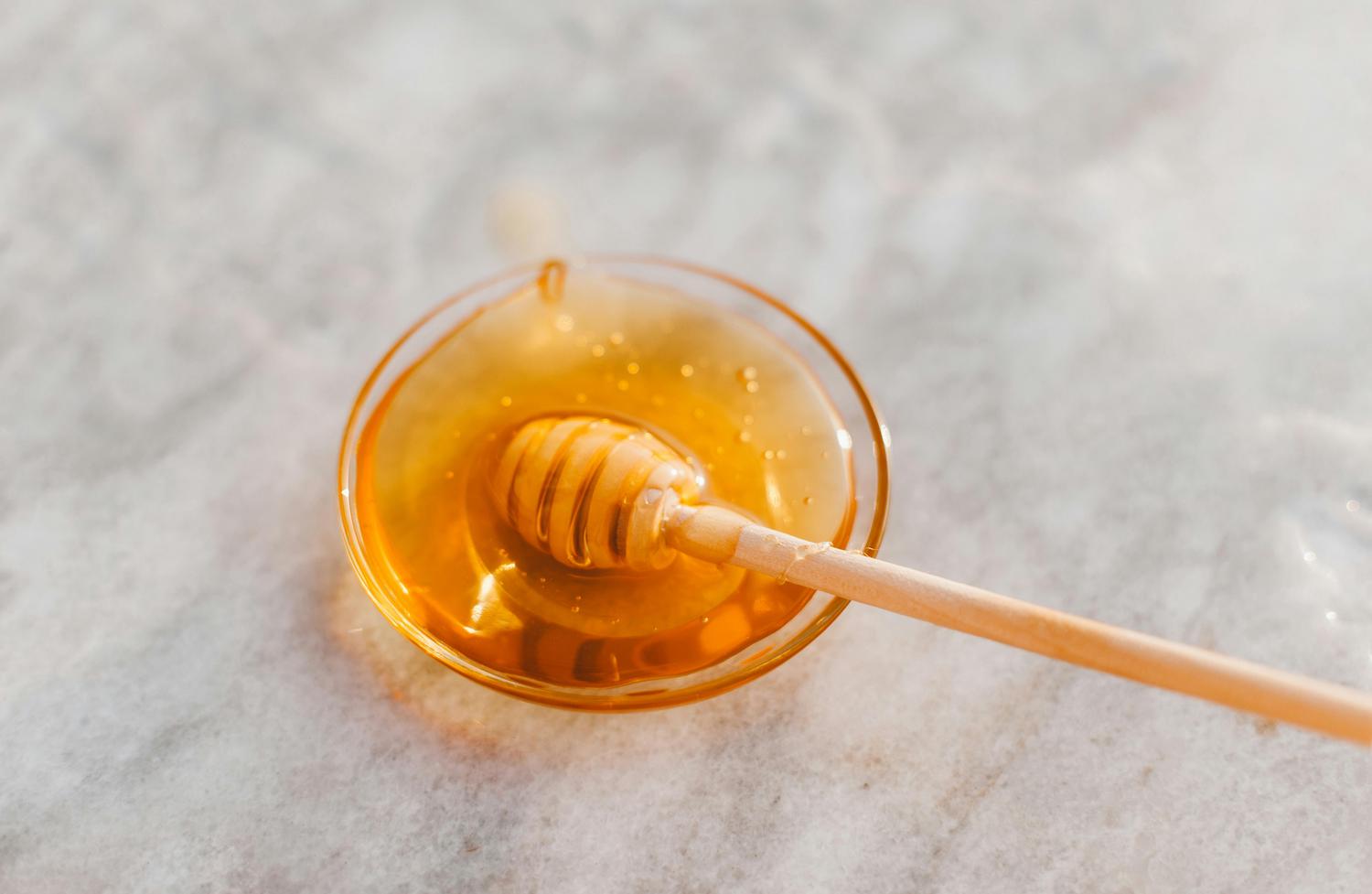 Golden honey fills a small glass bowl with a wooden honey dipper resting inside and extending to the right, all sitting on a textured white surface. The honey appears thick and viscous, with a few small bubbles visible.