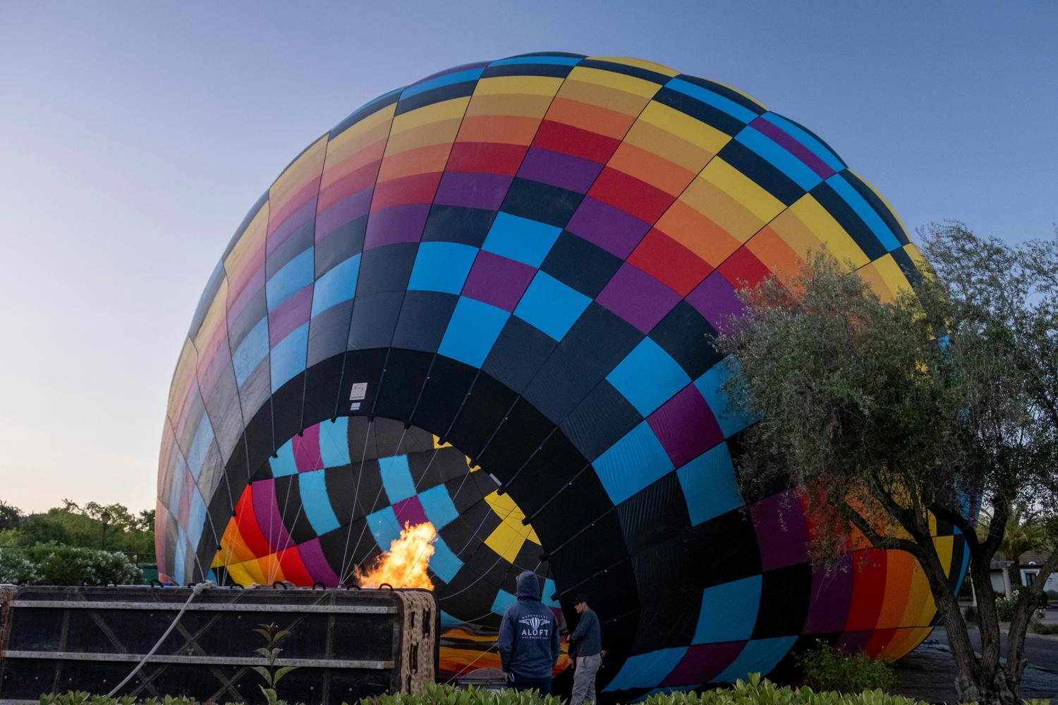 A partially inflated hot air balloon, brightly colored with squares in hues of yellow, orange, red, purple, and blue, is shown being filled with flames. Two figures stand near the flame source.