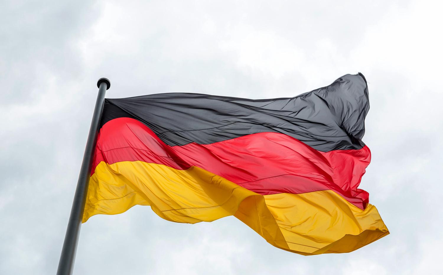 The German flag, featuring black, red, and yellow horizontal stripes, waves against a cloudy sky. The flag is attached to a dark metal pole.