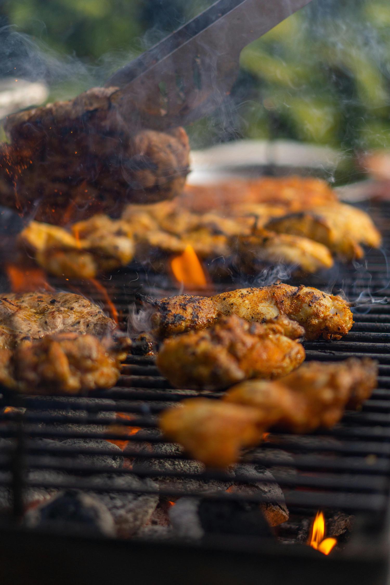 Seasoned chicken and other cuts of meat cook on a smoky barbecue grill with flames and glowing coals. A utensil turns a piece of meat at the top.