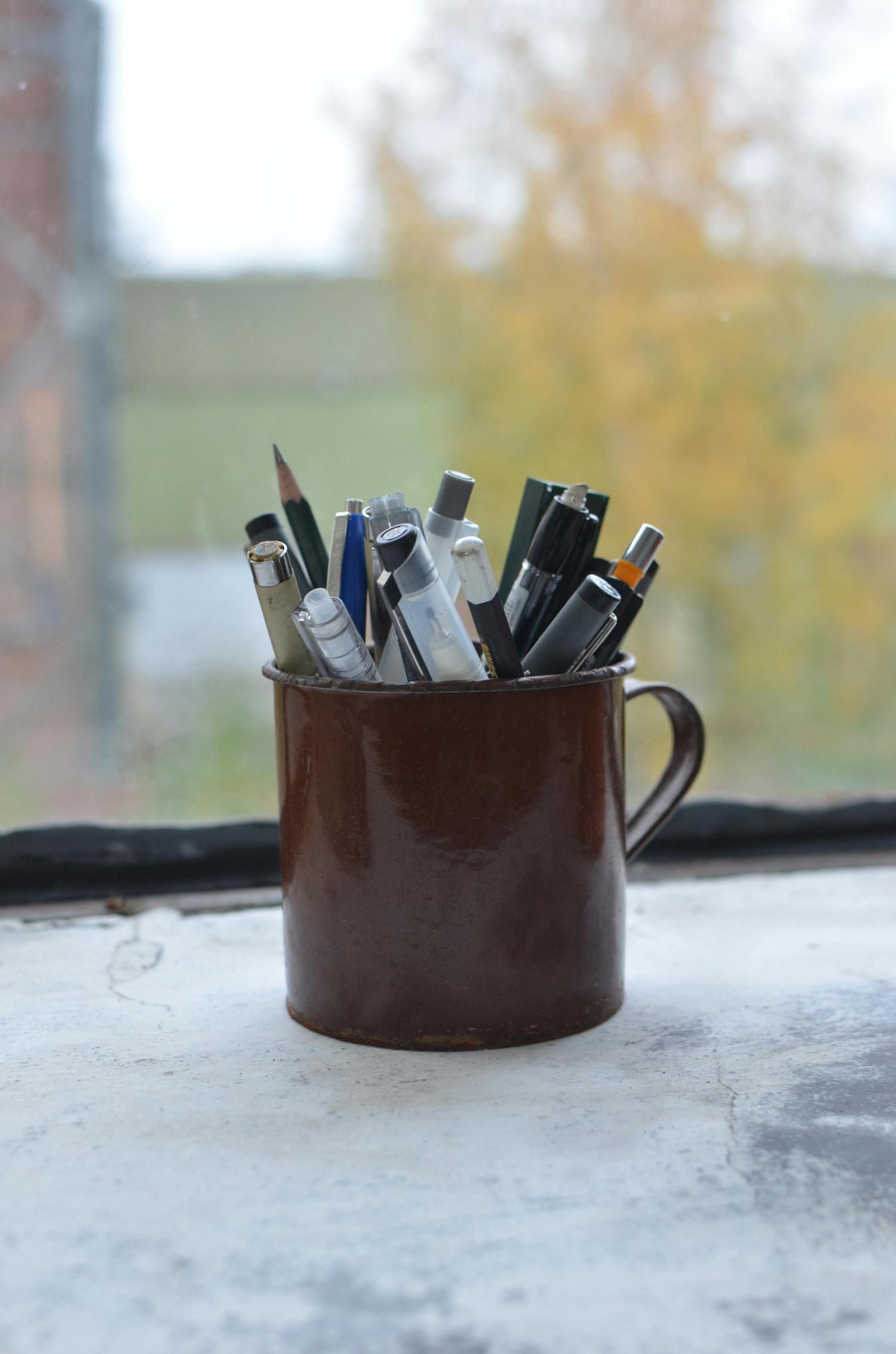 A rustic brown mug filled with an assortment of pens and pencils rests on a weathered windowsill, with a blurry green and yellow outdoor background.