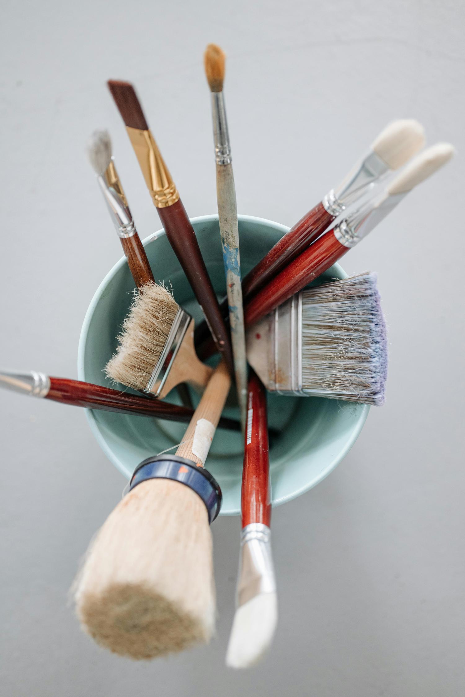 An overhead view shows numerous paintbrushes with varied handles and bristles, some stained with paint, gathered in a light blue container.