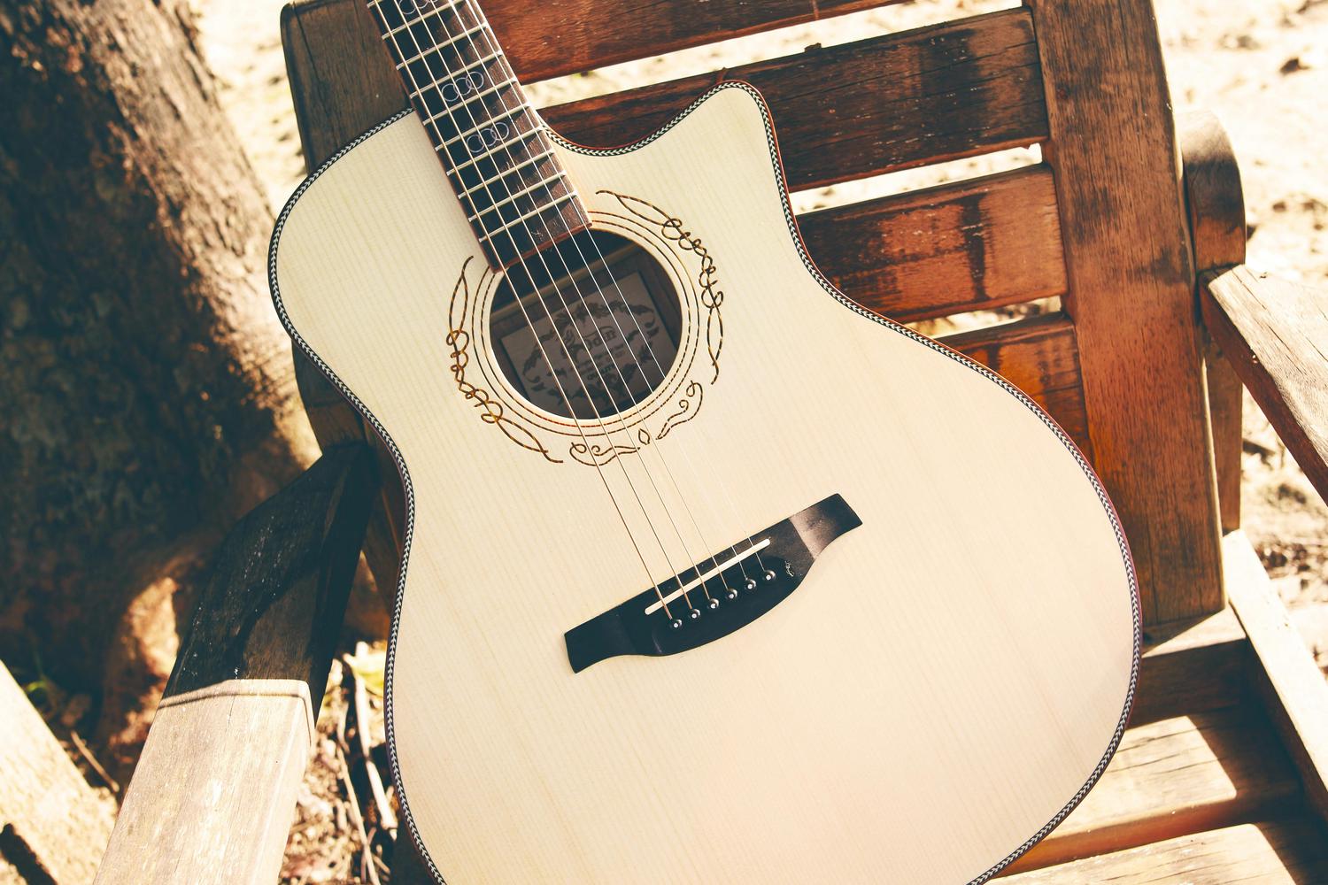 A light-colored acoustic guitar with decorative inlay around the soundhole and body edges rests on a wooden chair outdoors, with a tree trunk and sunlight in the background.